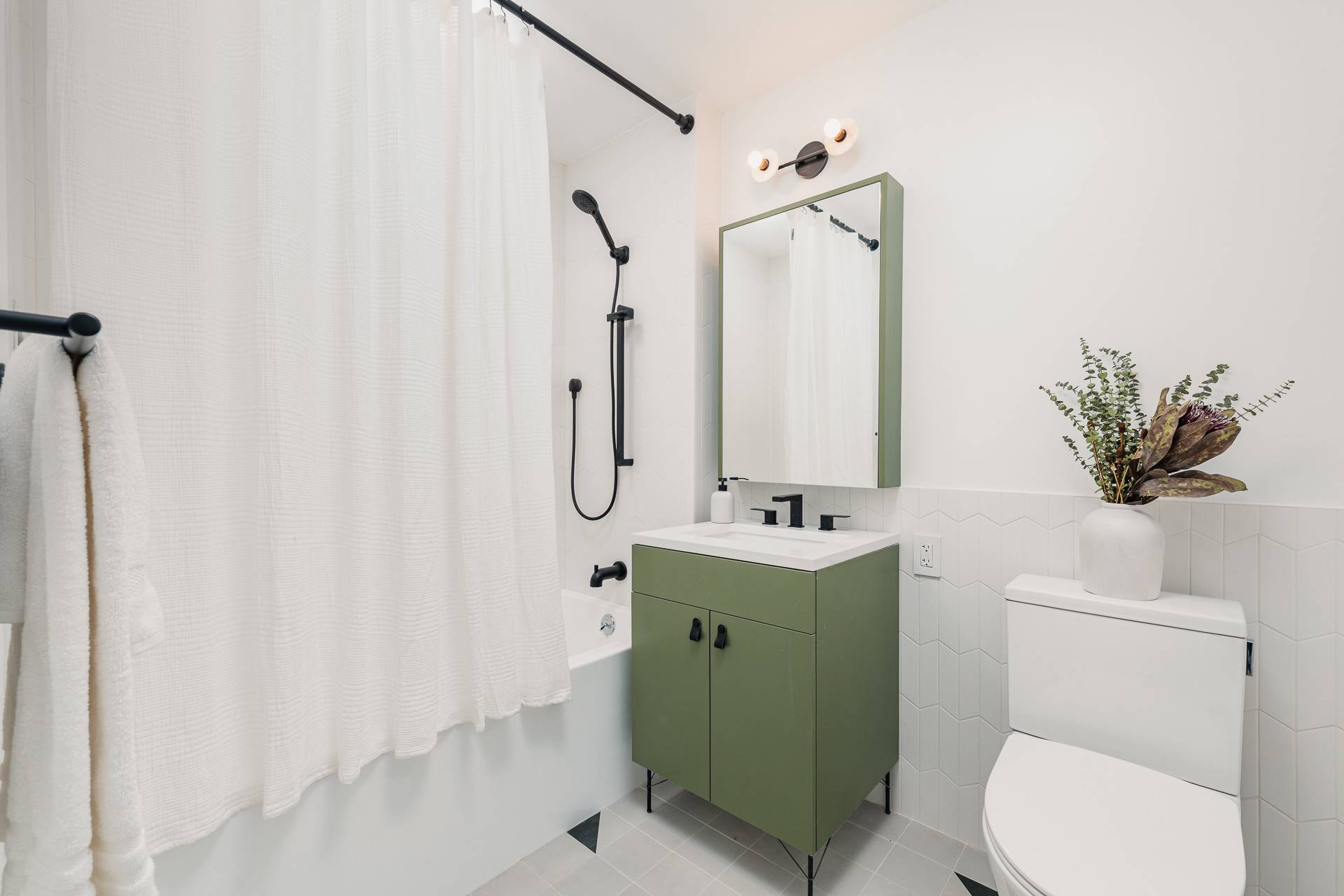 modern bathroom with light and white tiling on floors and bottom portion of wall, green tone vanity and mirror and shower tub with dark fixtures